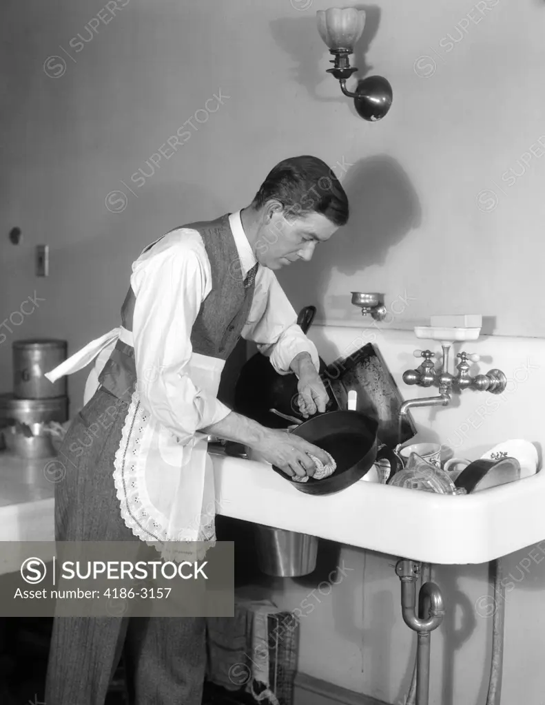 1930S Man Dressed In Shirt Tie Vest & Apron Bent Over A Sink Full Of Dishes  Cleaning An Iron Skillet With A Dishcloth - SuperStock