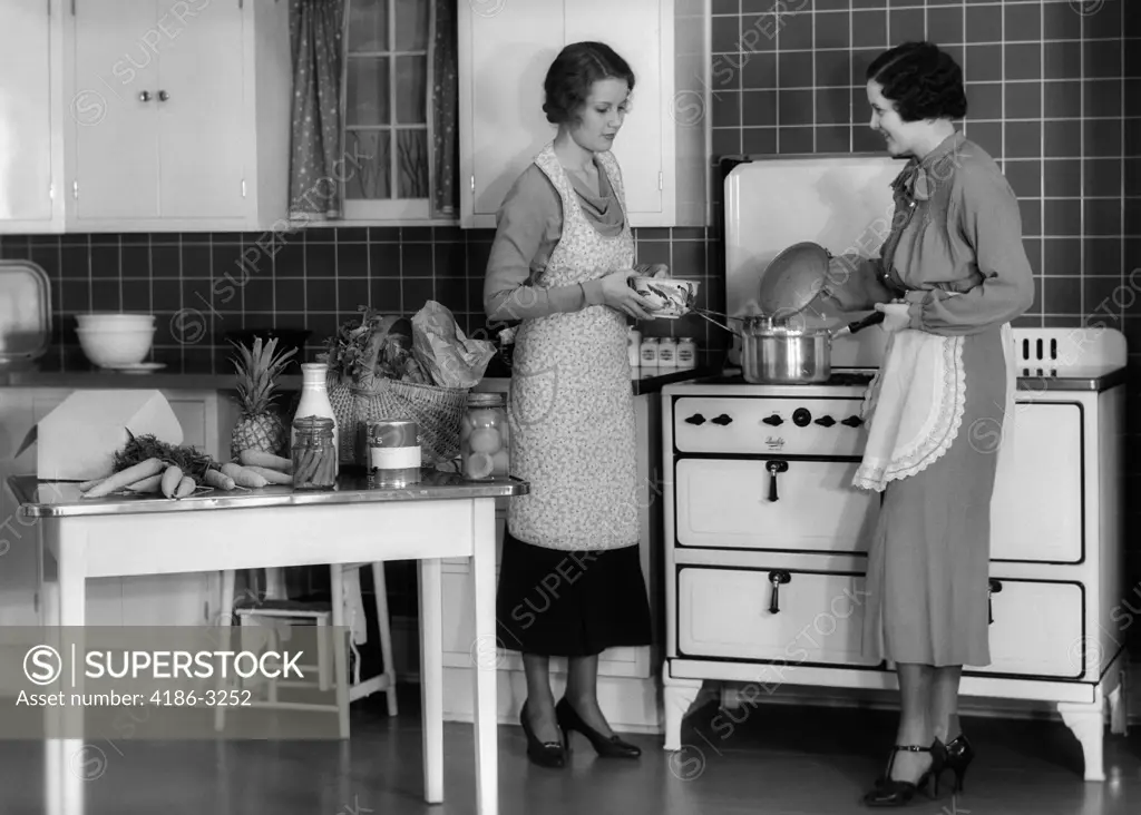 1930S Woman Housewife And Friend Wearing Apron Cooking Food In Kitchen On Gas Stove Indoor