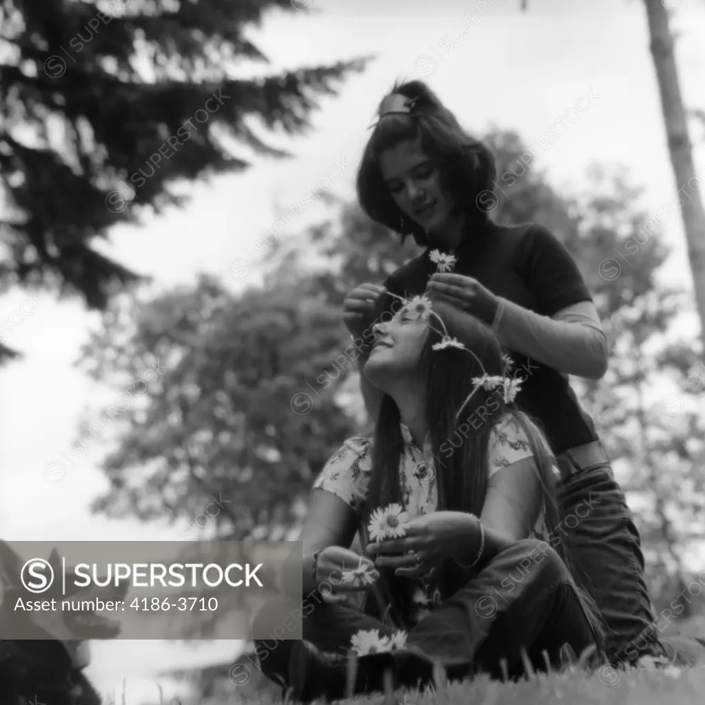 1970S Two Teen Hippie Girls Outdoors Sitting In Grass One Is Putting Flowers In Others Hair