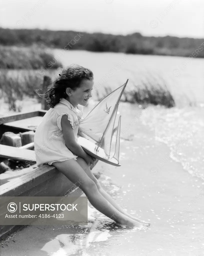 1930S Young Girl At Seashore Holding Sailboat Toy Sitting On Edge Of Rowboat Feet In Water