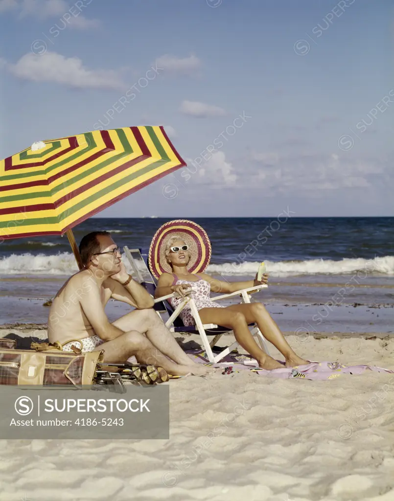 1950S 1960S Middle Age Couple Sitting On Beach Under Umbrella Man With Golf Clubs Looks Bored Wife Happy