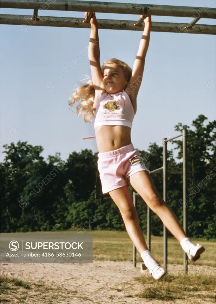 1970s GIRL HANGING FROM MONKEY BARS IN PLAYGROUND WEARING PINK HALTER TOP AND SHORTS