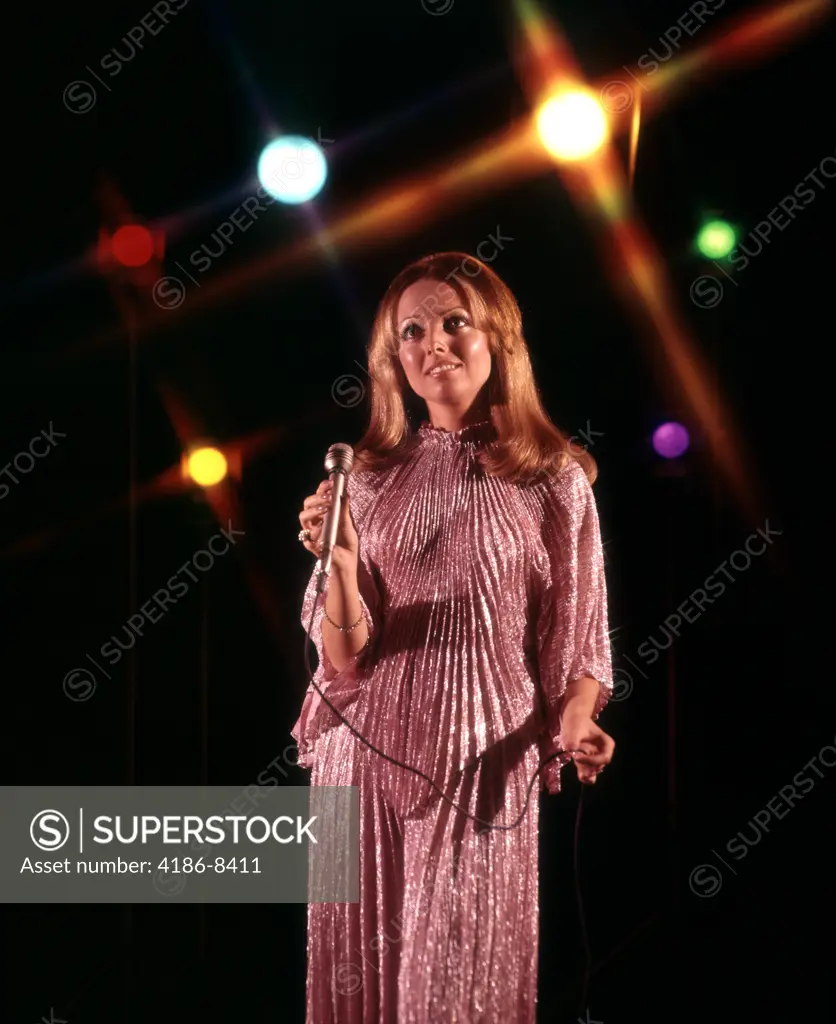 1970S Young Woman Entertainer With Long Hair Silver Pleated Dress And Holding Microphone Singing Among Stage Lights