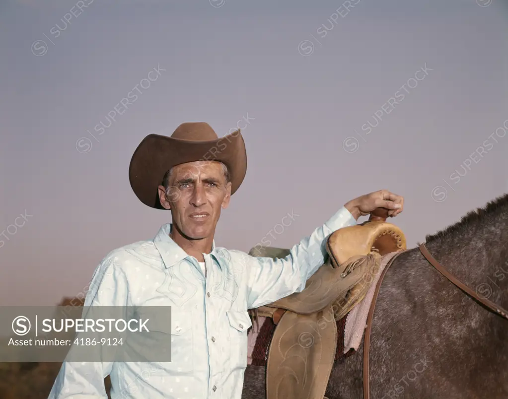 1960S Cowboy Leaning Against Saddle On Horse Wearing Cowboy Hat