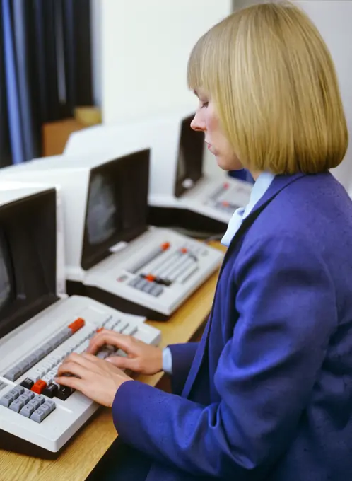 1980s BLOND WOMAN WEARING BLUE SUIT WORKING SITTING AT COMPUTER TERMINAL TYPING HANDS ON KEYBOARD