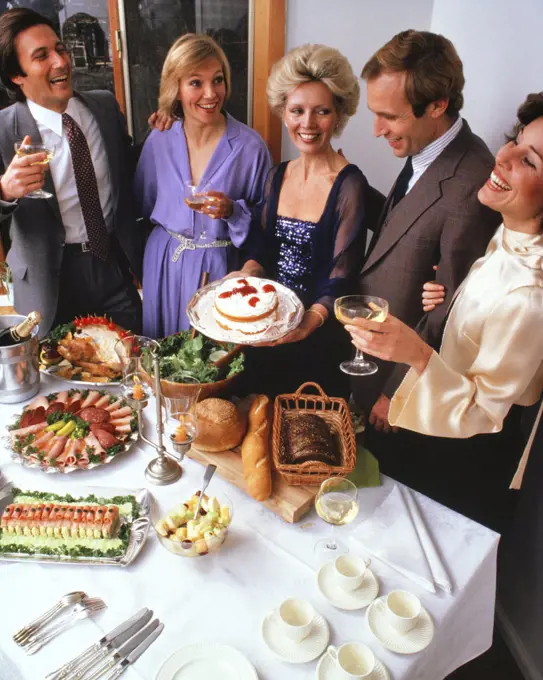 1980s MEN AND WOMEN TOASTING THEIR HOSTESS HOLDING STRAWBERRY SHORTCAKE DESSERT AT PARTY BUFFET TABLE