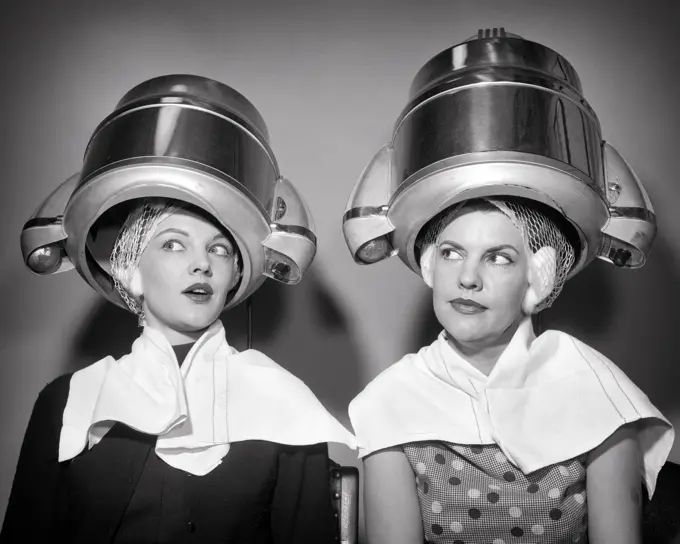 1950s TWO WOMEN SITTING UNDER BEAUTY SALON HAIR DRYERS WEARING HAIRNETS TOWELS TALKING GOSSIP