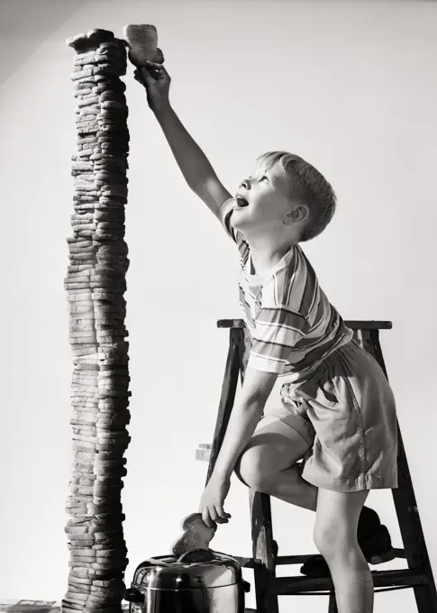 1950s EAGER BOY STANDING ON STEP LADDER REACHING THE TOP BUILDING A TOWER OF TOASTED BREAD SLICES