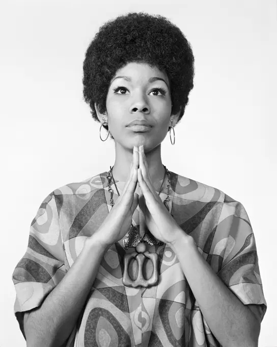 1960s AFRICAN AMERICAN WOMAN AFRO HAIRDO LOOKING AT CAMERA ARMS FOLDED IN PRAYER MEDITATION WEARING HOOP EARRINGS PRINT BLOUSE