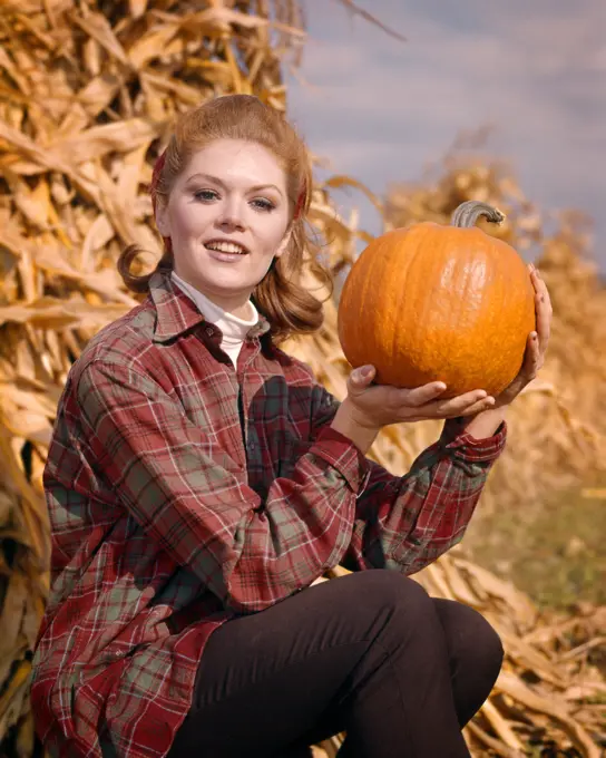 1960s SMILING RED HAIR WOMAN WEARING A PLAID SHIRT SITTING BY CORN SHOCK LOOKING AT CAMERA HOLDING UP PUMPKIN