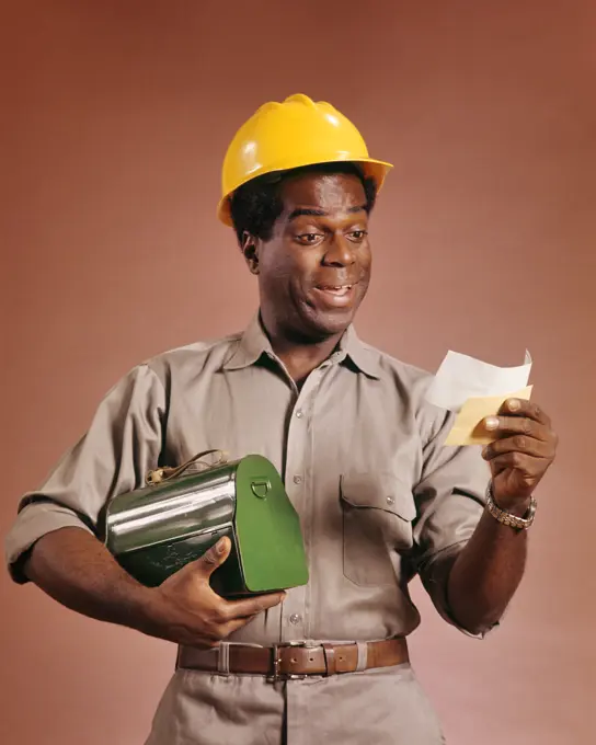 1970s AFRICAN AMERICAN WORKMAN HOLDING LUNCHBOX AND PAYCHECK SMILING WEARING A YELLOW HARD HAT