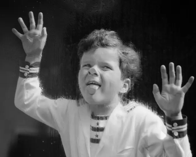 1930s ODD LAUGHING CURLY HAIR BOY WEARING NAUTICAL STYLE SHIRT PLACING HANDS AND STICKING TONGUE ON RAIN WET WINDOW GLASS PANE