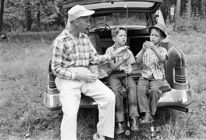 1960s MAN FATHER AND TWO BOYS SONS TWINS SITTING ON REAR BUMPER OF CAR PREPARING FISHING GEAR