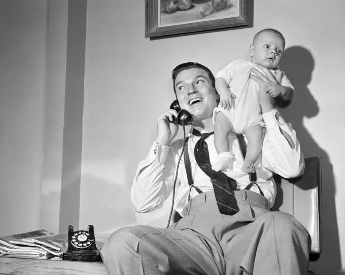 1940s 1950s MAN FATHER TALKING ON TELEPHONE HOLDING BABY BOY UP WITH ONE HAND