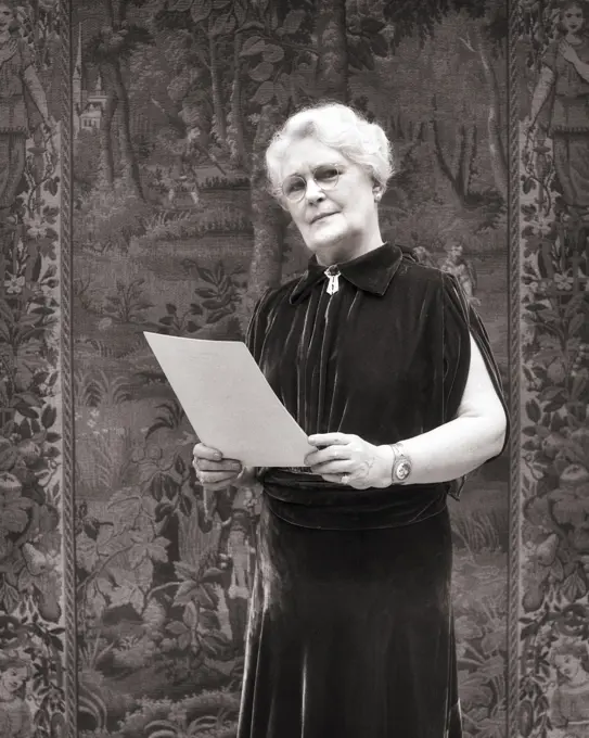 1930s elegant senior woman WEARING velvet dress holding paper GIVING A SPEECH STANDING BEFORE A TAPESTRY LOOKING AT CAMERA