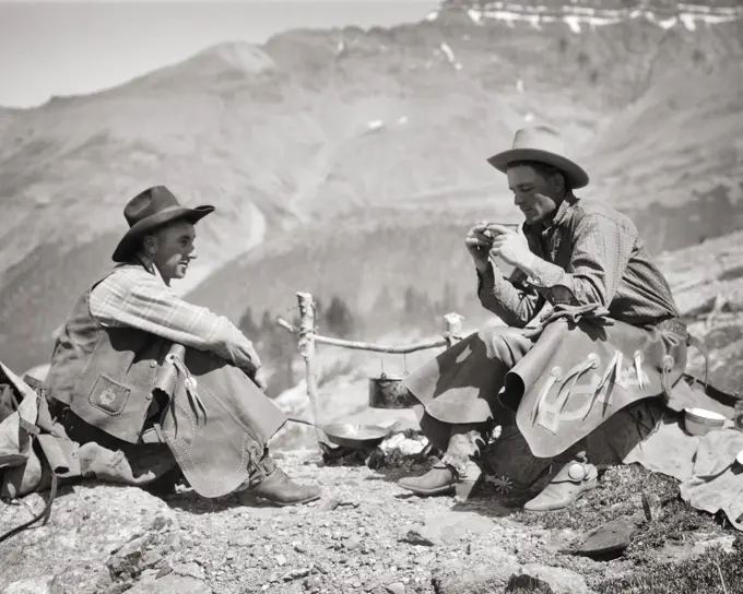 1920s 1930s TWO MEN WESTERN COWBOYS SITTING BY CAMPFIRE DRINKING COFFEE TALKING SUMMIT OF PASS NEAR BOW LAKE ALBERTA CANADA