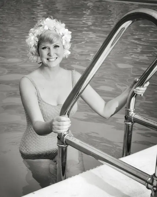 1960s BLOND WOMAN IN ONE PIECE BATHING SUIT AND FLOWER STYLE SWIM CAP CLIMBING OUT OF SWIMMING POOL SMILING LOOKING AT CAMERA