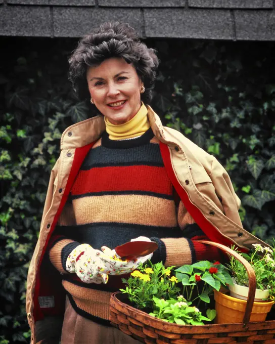 1980s SMILING WOMAN WEARING SWEATER JACKET YELLOW TURTLENECK GARDENING GLOVES HOLDING BASKET OF PLANTINGS LOOKING AT CAMERA