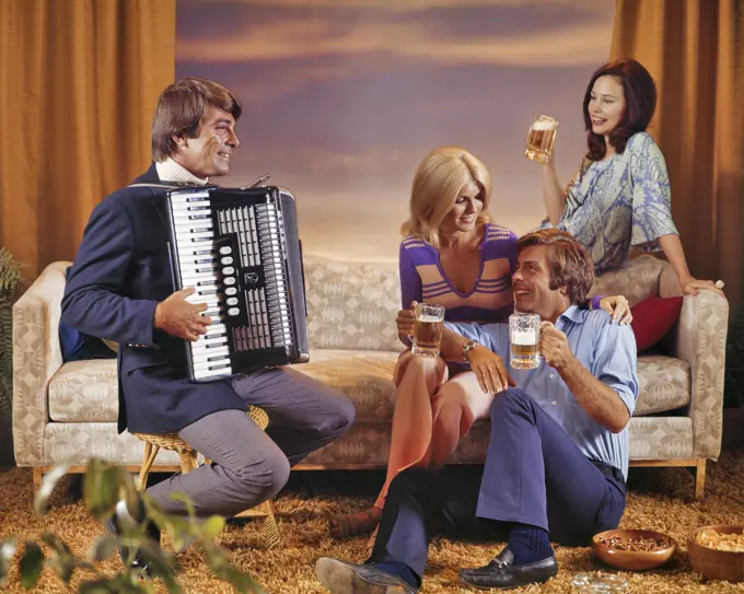 1970s 1980s TWO SMILING COUPLES MEN AND WOMEN INFORMALLY SOCIALIZING THREE DRINKING BEER AND ONE MAN PLAYING AN ACCORDION