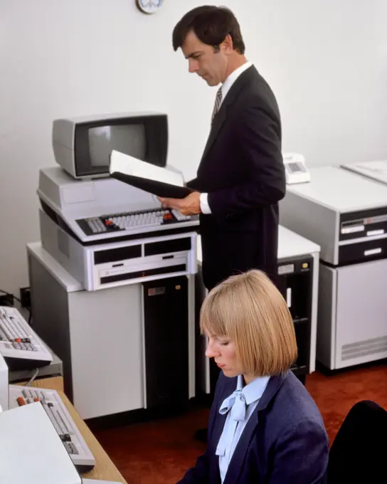 1980s OFFICE WITH EARLY COMPUTER HARDWARE WOMAN SEATED AT TERMINAL KEYBOARD MAN STANDING READING MANUAL