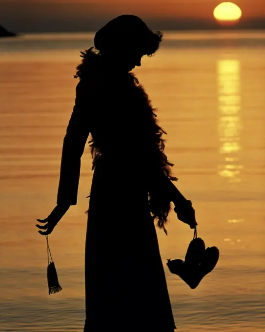 1970s ANONYMOUS SILHOUETTE PROFILE OF YOUNG WOMAN WEARING FEATHER BOA CARRYING EVENING PURSE AND SHOES WALKING BY MOONLIT WATER