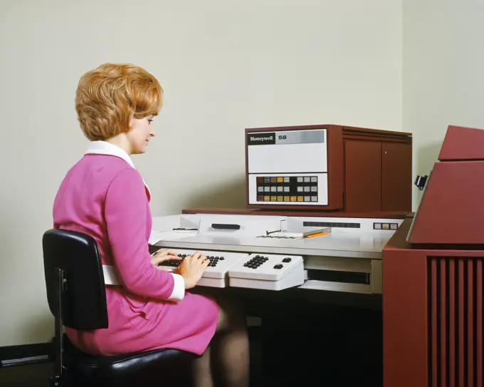 1970s WOMAN SITTING AT KEYBOARD OPERATING A HONEYWELL 58 ELECTRONIC DATA PROCESSING MACHINE