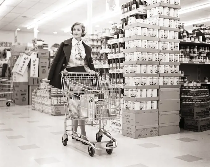 1960s 1970s WOMAN COAT DRAPED ON SHOULDERS PUSHING SHOPPING CART IN GROCERY STORE SUPERMARKET