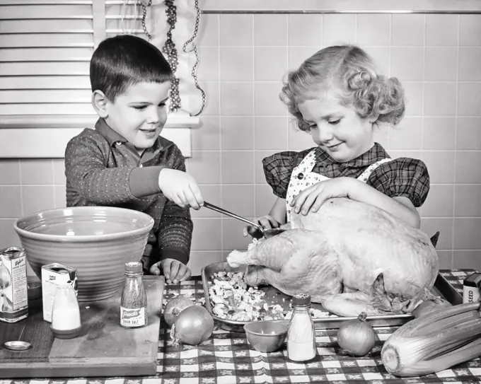 1950s SMILING BOY AND GIRL BROTHER AND OLDER SISTER LADLING BREAD STUFFING INTO THANKSGIVING TURKEY ON KITCHEN TABLE