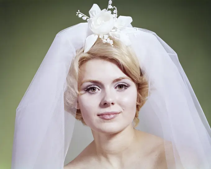 1960s PORTRAIT OF BLONDE BRIDE LOOKING AT CAMERA  WEARING NET VEIL WITH WHITE FLORAL ACCESSORY