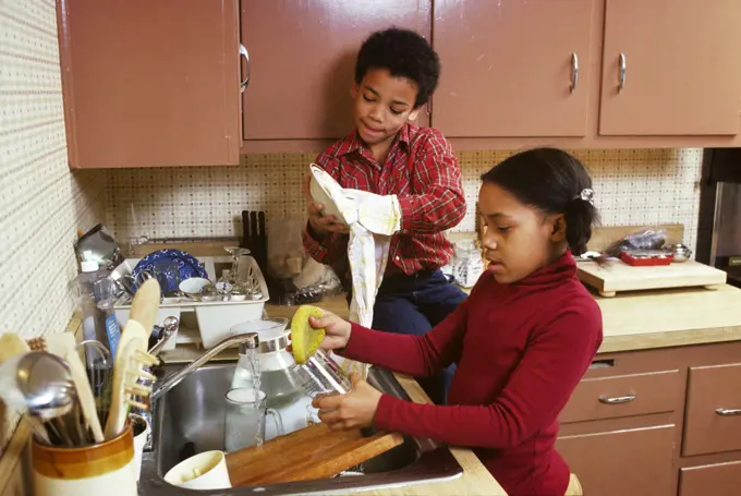1970s 1980s AFRICAN AMERICAN BOY AND GIRL BROTHER AND SISTER TOGETHER WASHING DISHES BY HAND BOY SITTING ON KITCHEN COUNTER