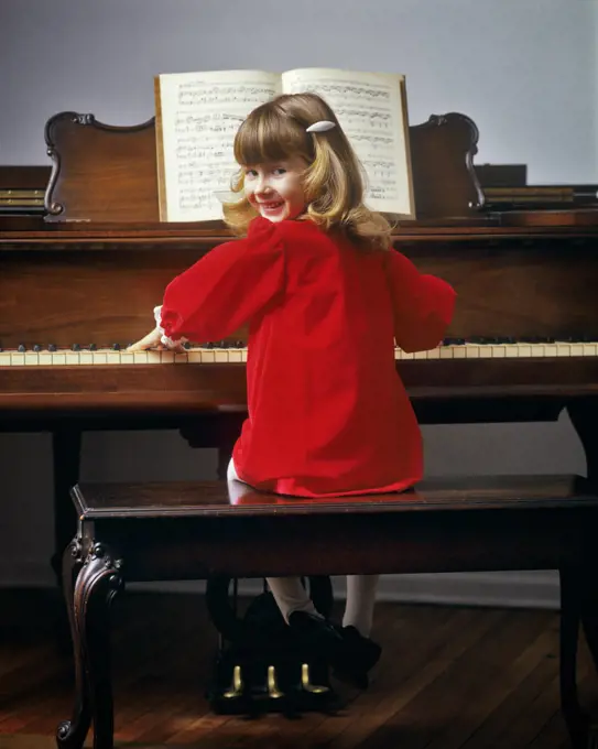 1980s CUTE SMILING YOUNG BLOND GIRL WEARING READ DRESS SITTING PLAYING PIANO TURNING HEAD OVER SHOULDER LOOKING AT CAMERA