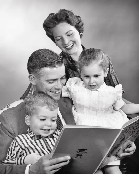 1950s FAMILY AT HOME WOMAN MAN GIRL BOY SMILING MOTHER WATCHING LOVING FATHER READ BOOK TO DAUGHTER AND SON