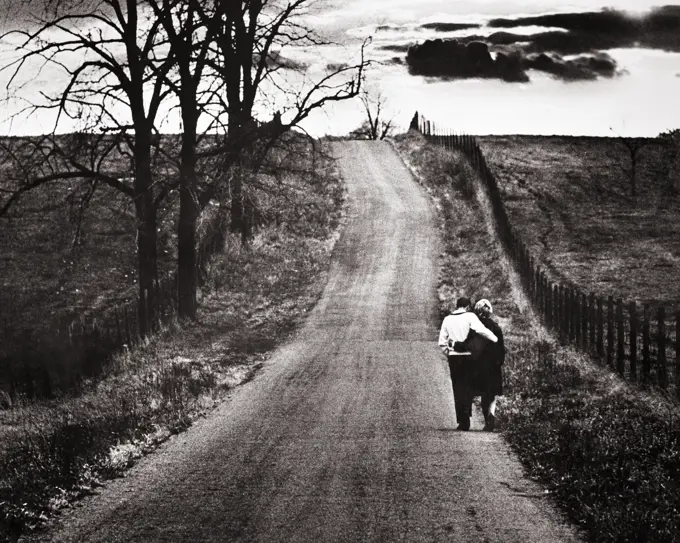 1960s BACK VIEW MATURE COUPLE ARM IN ARM WALKING DOWN LONELY ROAD BARREN AUTUMN LANDSCAPE WITH OMINOUS CLOUDS ON THE HORIZON