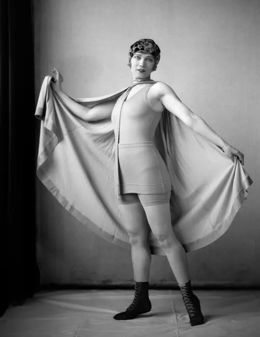 1920S Woman In Old-Fashioned Bathing Suit & Cap Holding Out Large Cape Tied Around Neck Looking At Camera