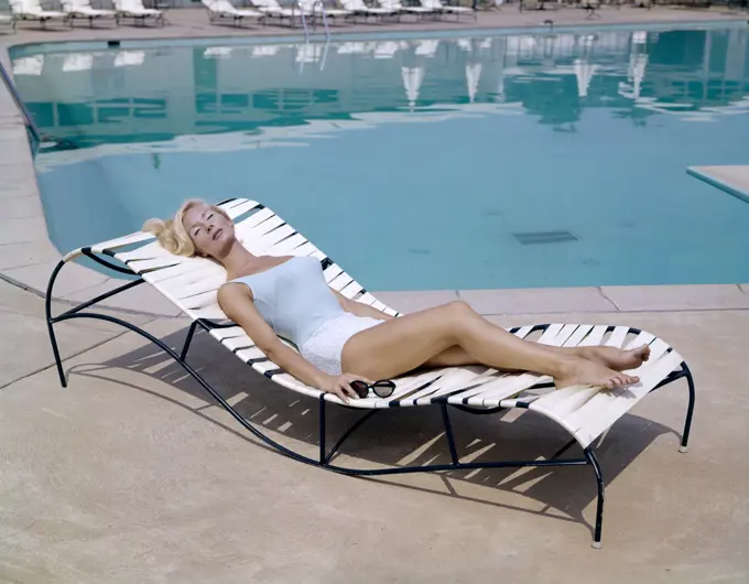 1960S Elegant Tall Blond Woman In Bathing Suit Reclining On A Lounge Chair By Swimming Pool Tanning In The Sunshine