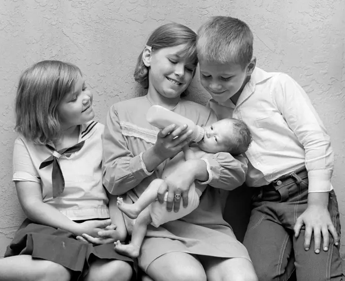 1970S Girl Feeding Bottle To Baby Sister With Brother & Sister Sitting On Either Side Of Her Looking On