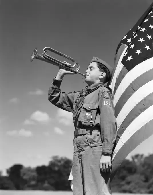 1950S Side View Of Boy Scout Standing In Front Of American Flag Bugling