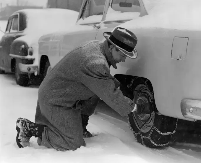 1950S Man In Coat & Hat Kneeling Down In Snow To Adjust Chains On Tires