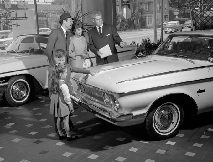 1960S Family Of 4 In Auto Showroom With Salesman Looking At White Plymouth