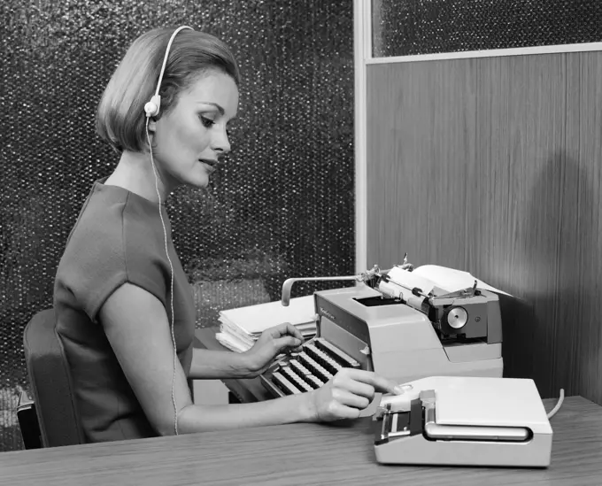 1960S Side View Of Secretary At Desk Behind Typewriter Wearing Headset & Turning Knob On Dictaphone