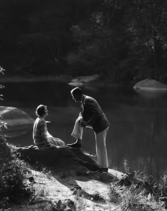 1920S 1930S Woman Sitting On Rock Lakeside Talking To Man Standing With Foot On Rock Leaning On Knee Listening