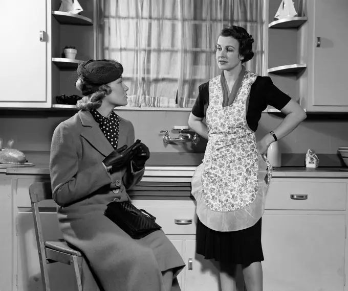 1930S 1940S Two Women Sitting In Kitchen Talking One Housewife Homemaker In Apron One In Coat And Hat Visiting Together Gossip