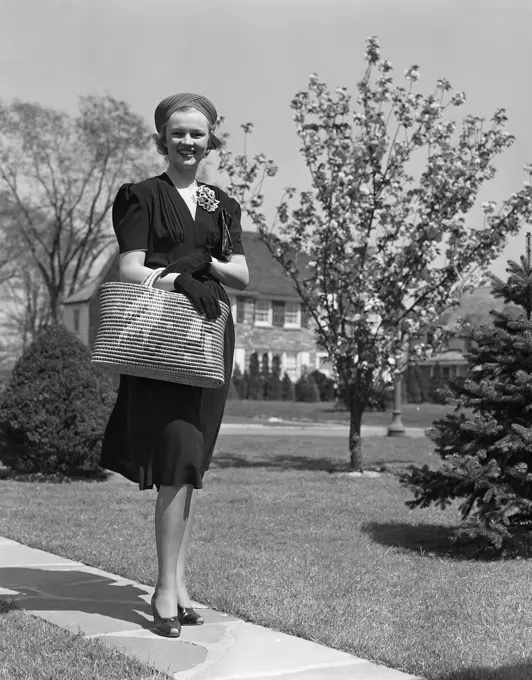 1930S 1940S Stylishly Dressed Woman Walking Down Suburban Street Carrying Straw Bag  