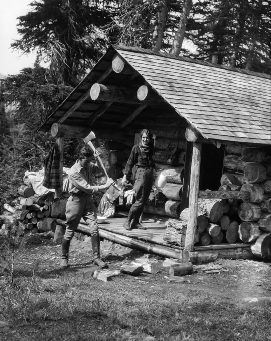 1920S 1930S Couple Woman Standing On Porch Of Log Cabin Holding Pitcher Man Chopping Wood Assiniboine Canada