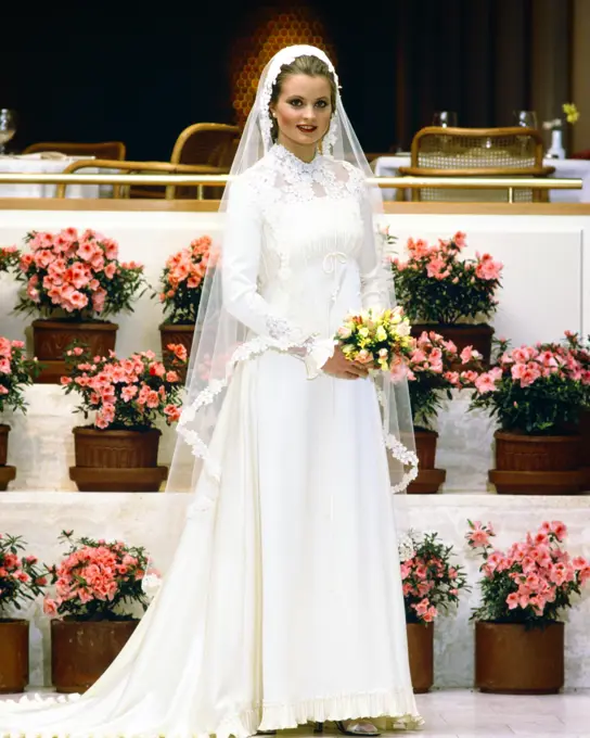 1970s 1980s SMILING BRIDE IN GOWN LOOKING AT CAMERA HOLDING YELLOW BOUQUET LONG VEIL LACE TRIM STANDING BY TIERS OF PINK AZALEAS