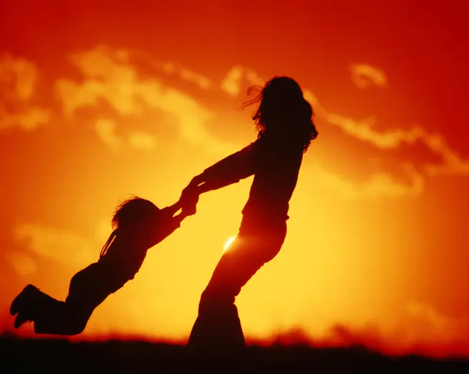 1970s 1980s AN ANONYMOUS SILHOUETTED JOYOUS MOTHER SWINGING CHILD GIRL OR BOY BY BOTH ARMS IN GOLDEN LIGHT SUNRISE OR SUNSET 