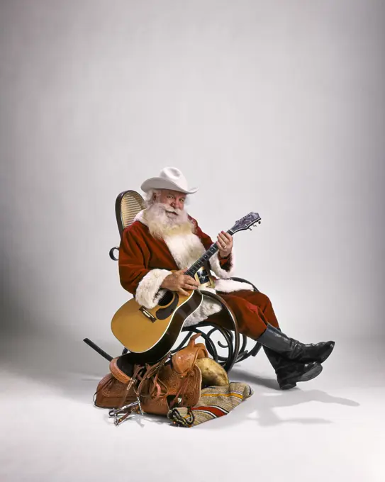 1980s COWBOY SANTA CLAUS LOOKING AT CAMERA SITTING IN BENTWOOD ROCKER BESIDE WESTERN SADDLE PLAYING GUITAR WEARING WHITE HAT