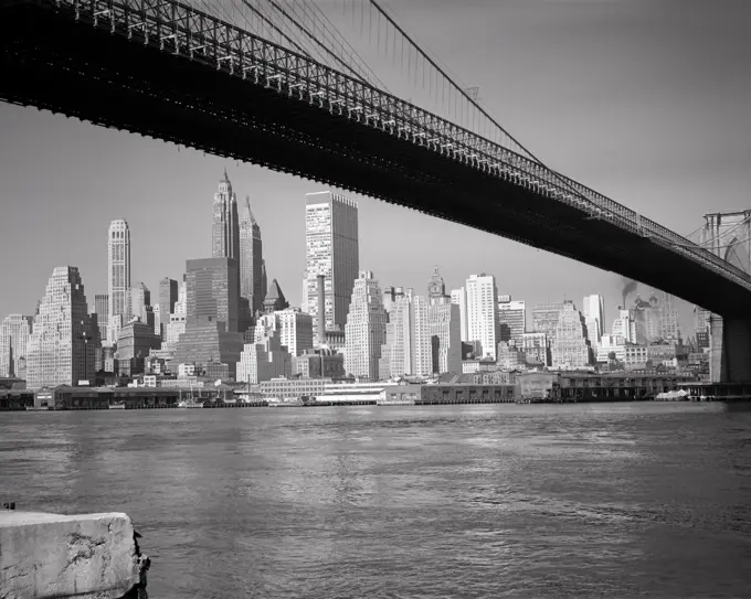 1960s DOWNTOWN FINANCIAL DISTRICT NEW YORK CITY FROM UNDER BROOKLYN BRIDGE OVER THE EAST RIVER