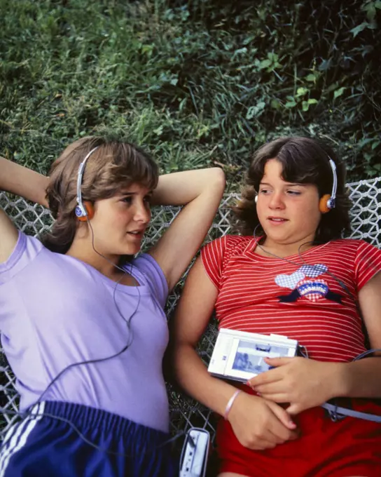 1980s TWO TEEN GIRLS LYING ON HAMMOCK LISTENING FOAM HEADPHONES TO SOUNDS MUSIC RECORDED CASSETTE ON PORTABLE MEDIA PLAYER 
