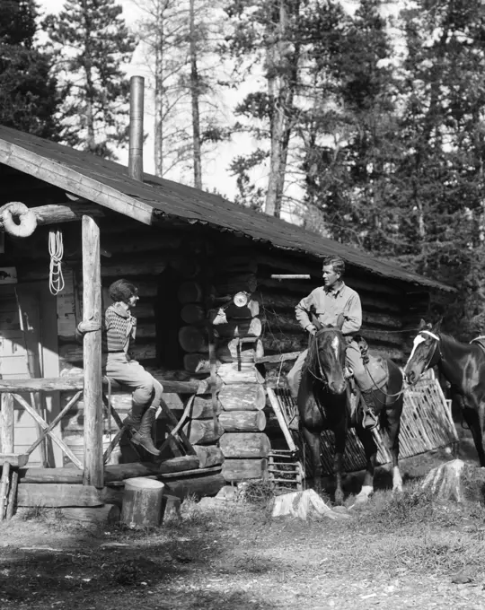 1920S 1930S Couple In Front Of Log Cabin Woman Sitting On Porch Railing Man On Horse Alberta Canada